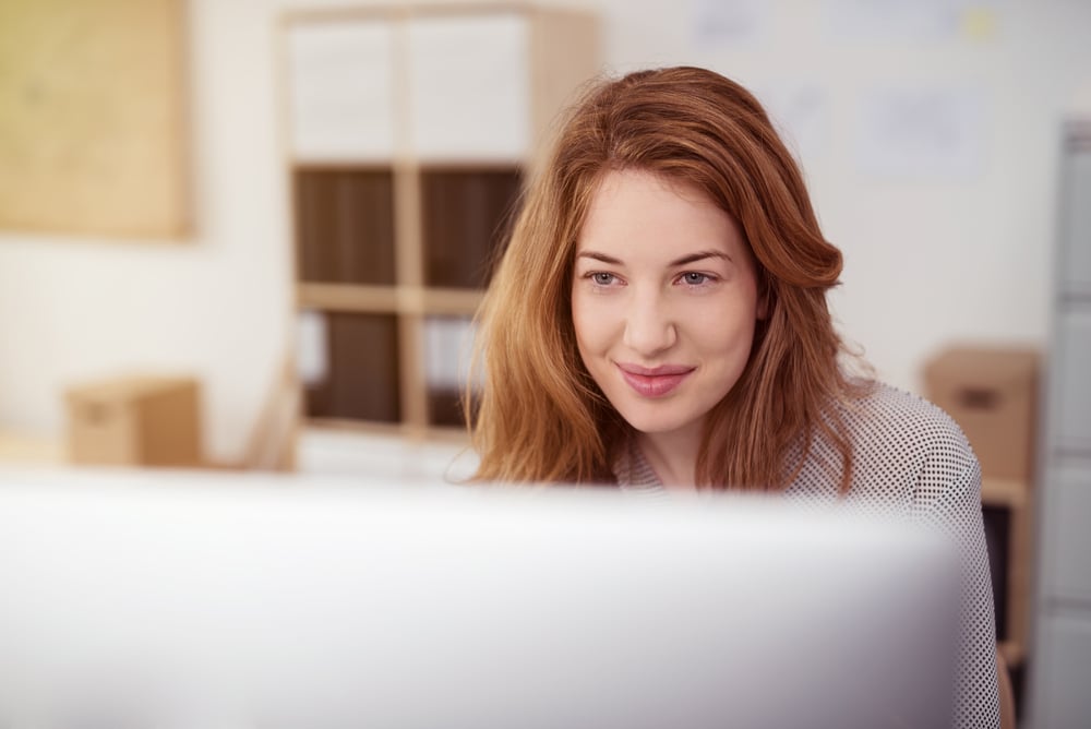 Attractive young woman working on a desktop computer smiling as she leans forwards using Rebuy for Shopify, view over the monitor