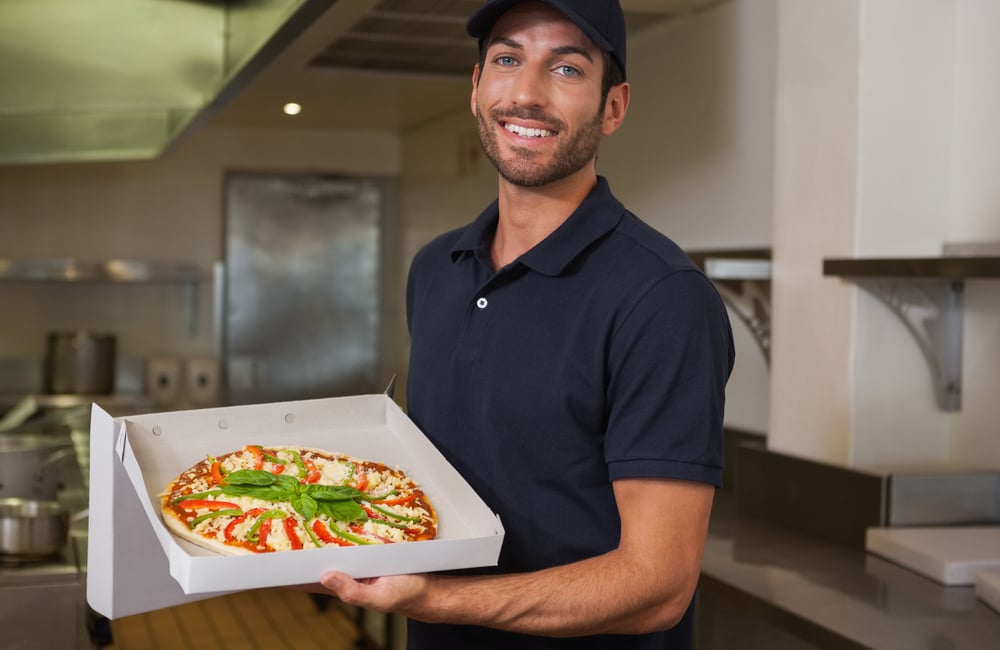 Happy pizza delivery man showing fresh pizza in a commercial kitchen